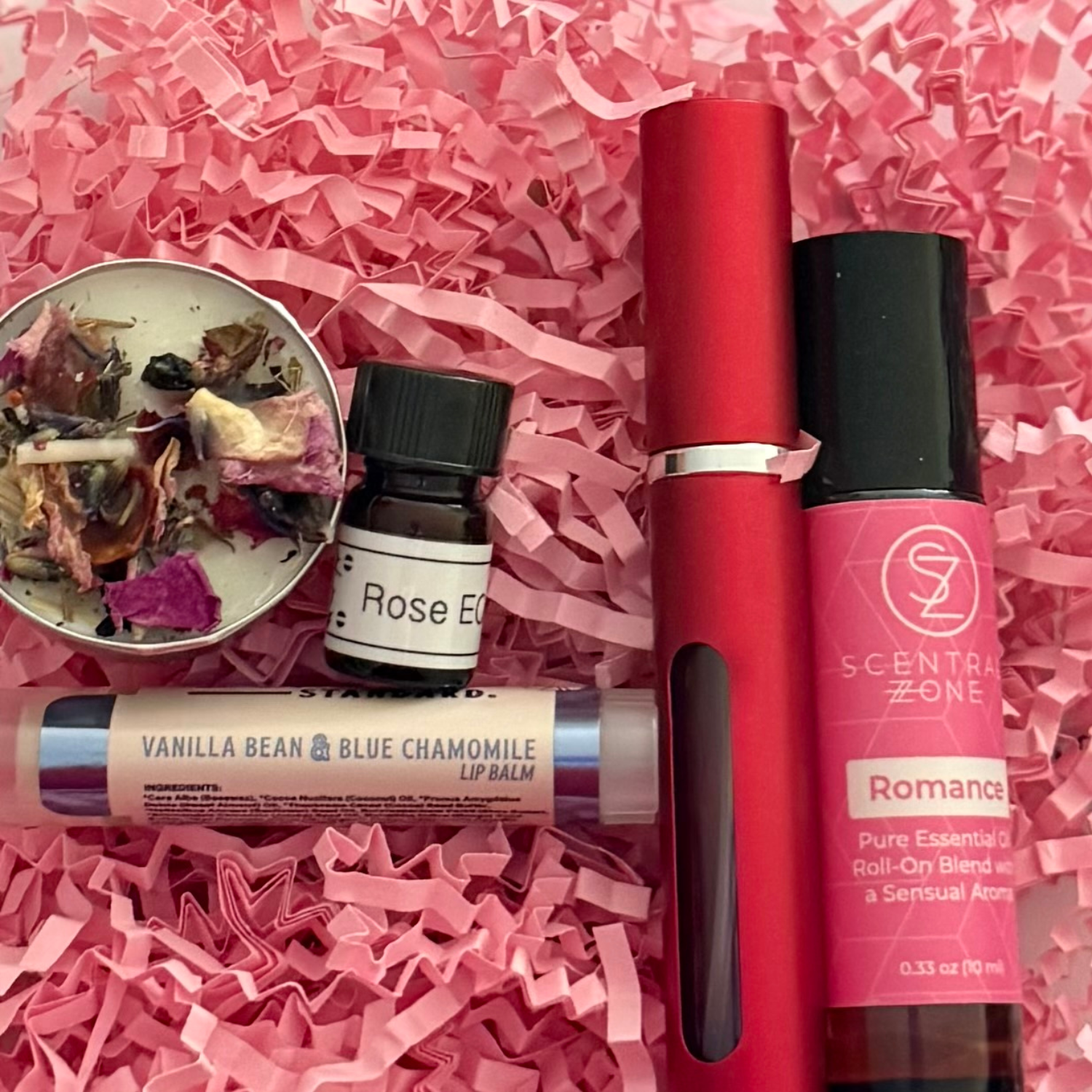 Collection of beauty products including a red roller, lip balm, and essential oil on pink shredded paper.